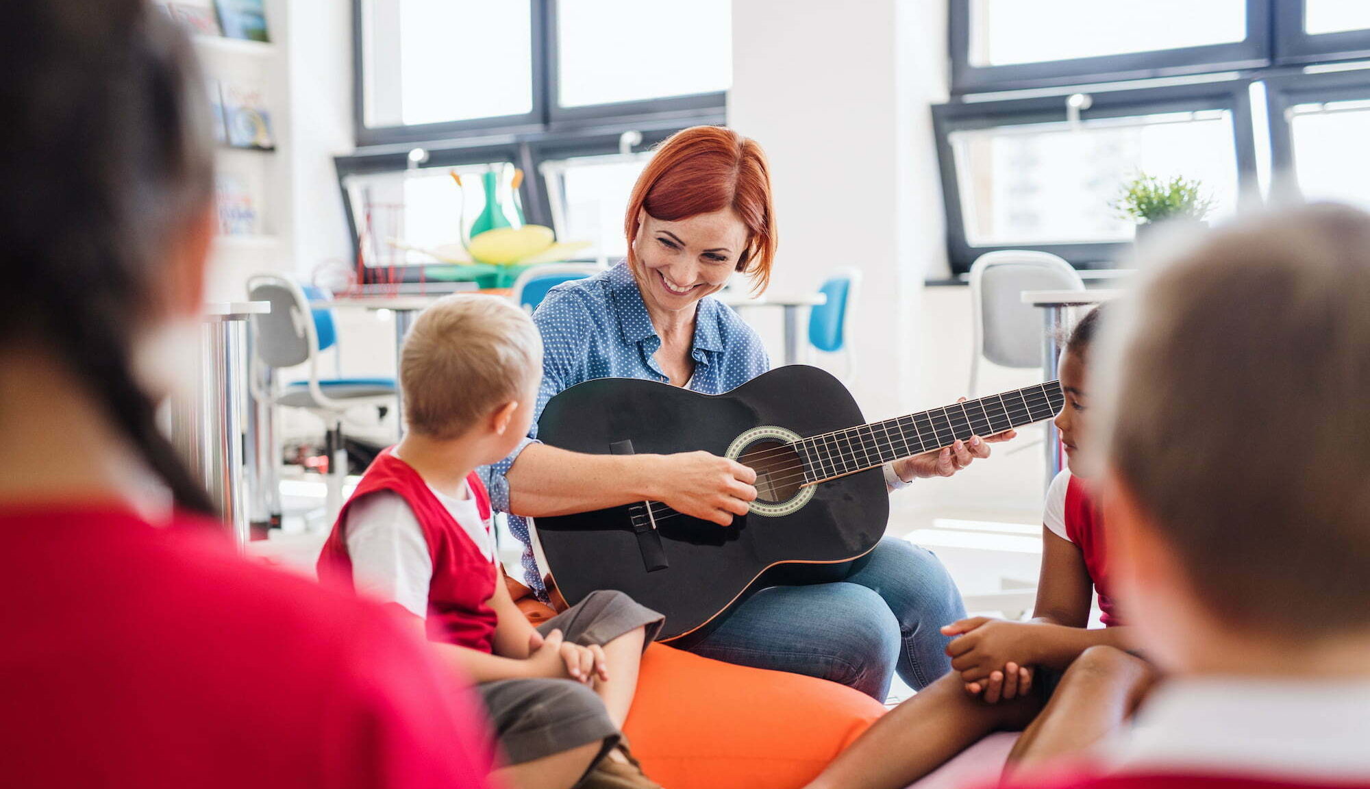 school kids and teacher with guitar sitting on the floor in class.