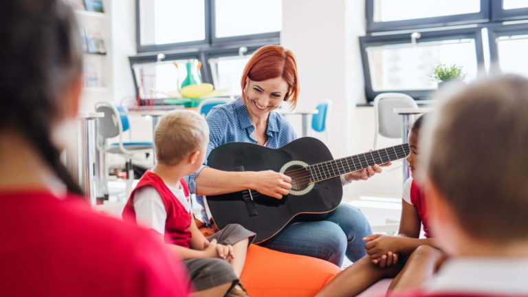 school kids and teacher with guitar sitting on the floor in class.